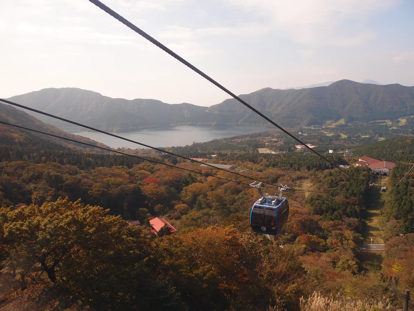 A blue gondola on its cable above autumn woods, with Lake Ashi and a ring of mountains ahead
