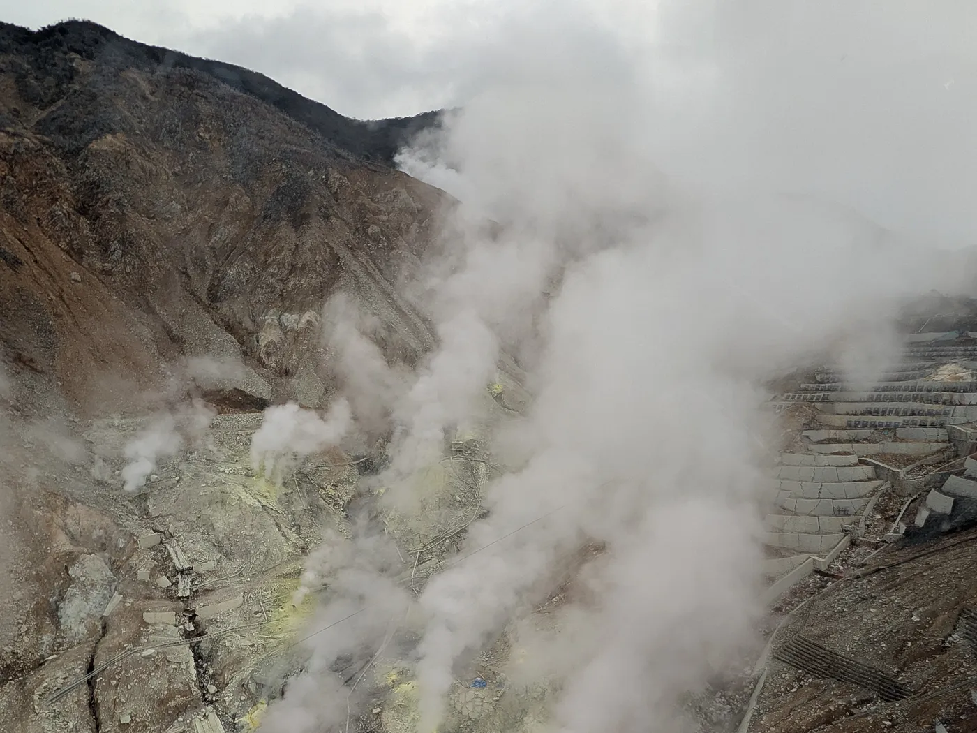 A steaming volcanic valley below a bare brown ridge, with white vapour rising from yellow sulphur vents