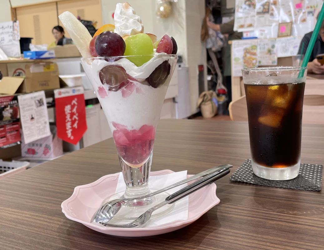 A tall glass parfait with grapes, peach and cream beside an iced coffee on a wooden table